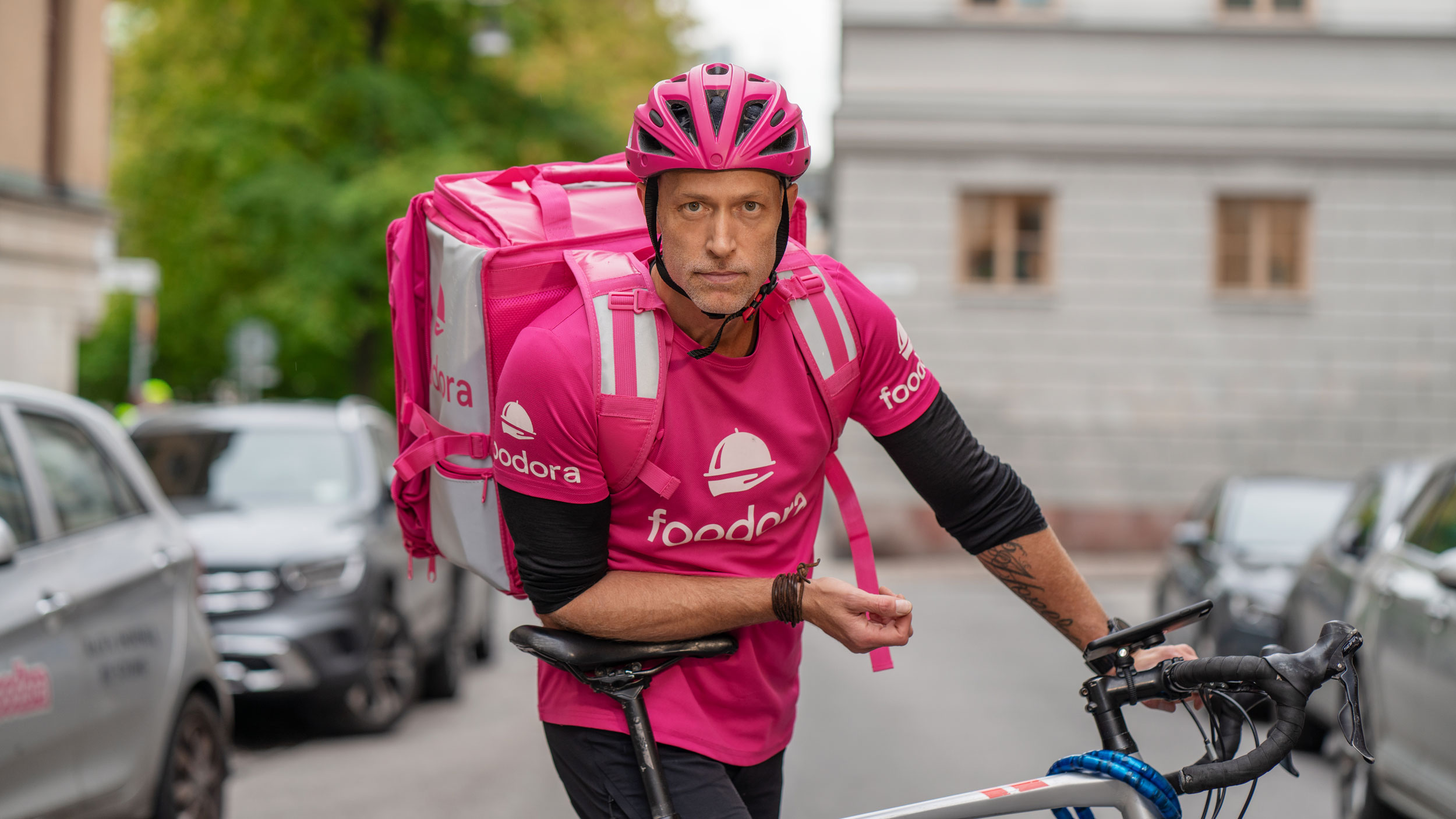En manlig Foodora-rider som hänger lutandes mot sin cykelsadel- iklädd sina rosa arbetskläder.